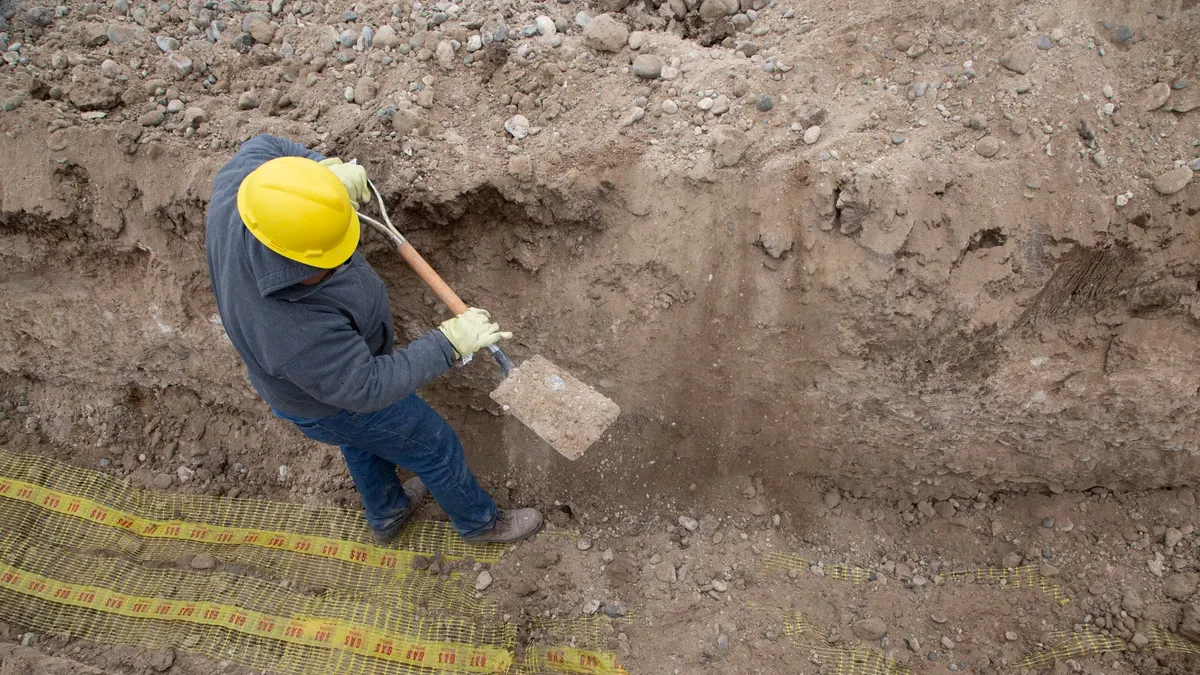 Worker in a hard hat digging a sewer trench beside yellow safety mesh